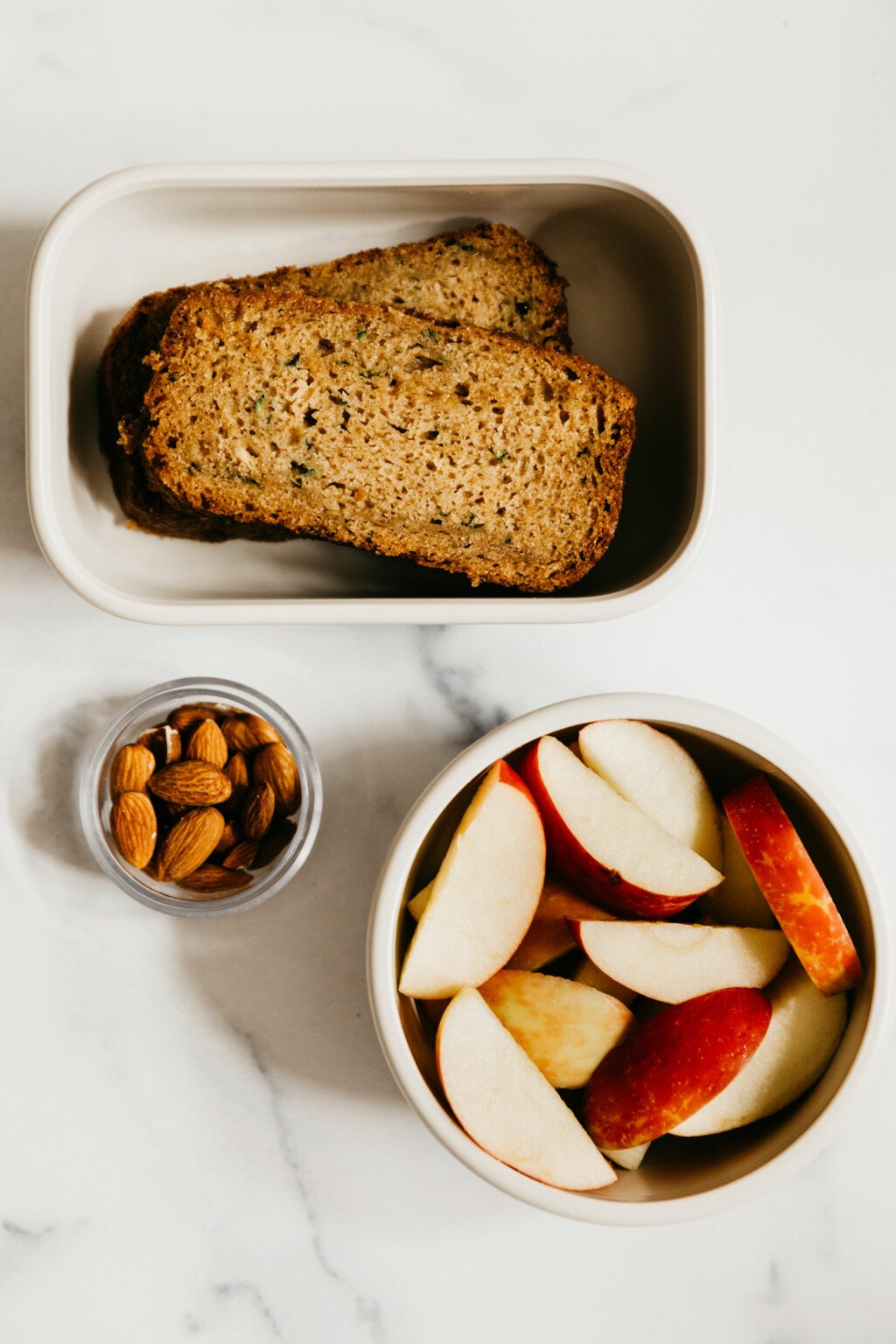 Two slices of zucchini bread, some apple slices, and raw almonds are packed in containers and resting on a white surface.