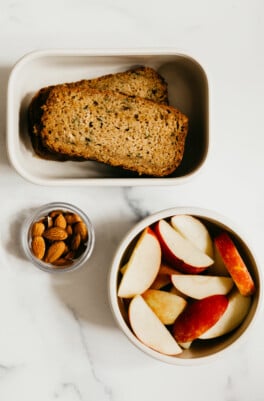 Two slices of zucchini bread, some apple slices, and raw almonds are packed in containers and resting on a white surface.