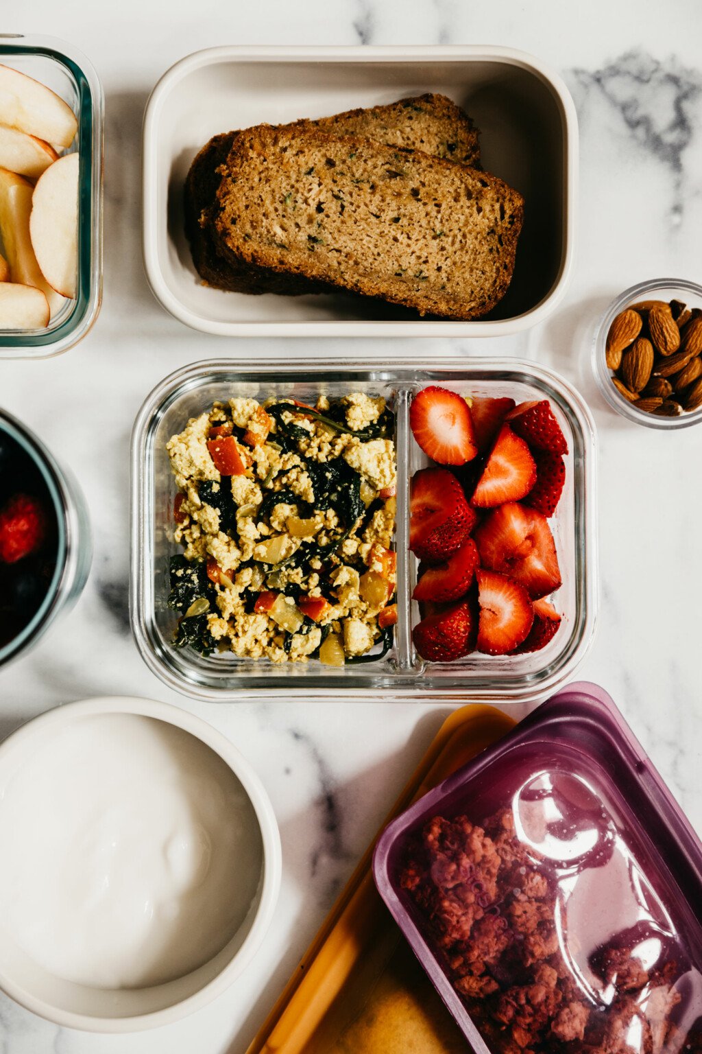 A few vegan meal prep breakfast ideas are laid out on a white marble surface, including tofu scramble, zucchini bread, yogurt, and more.