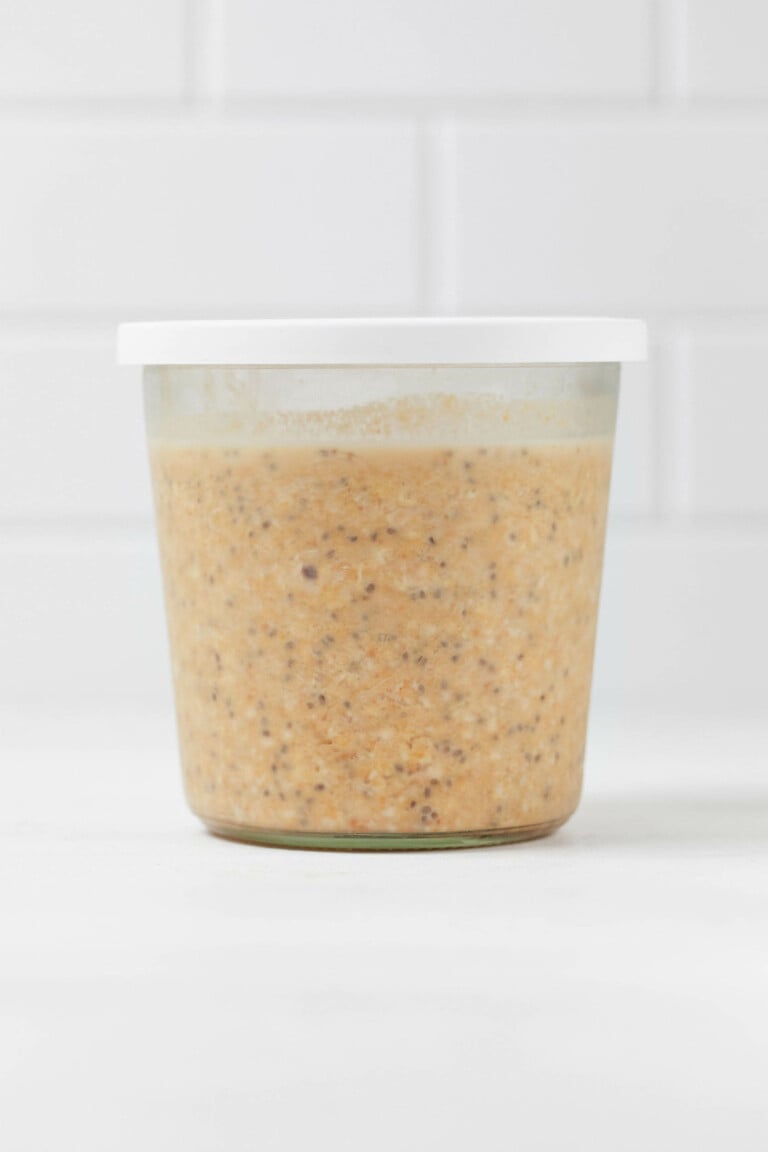 A glass mason jar with a white lid is resting on a white surface. It holds a mixture of vegan pumpkin overnight oats with chia seeds.