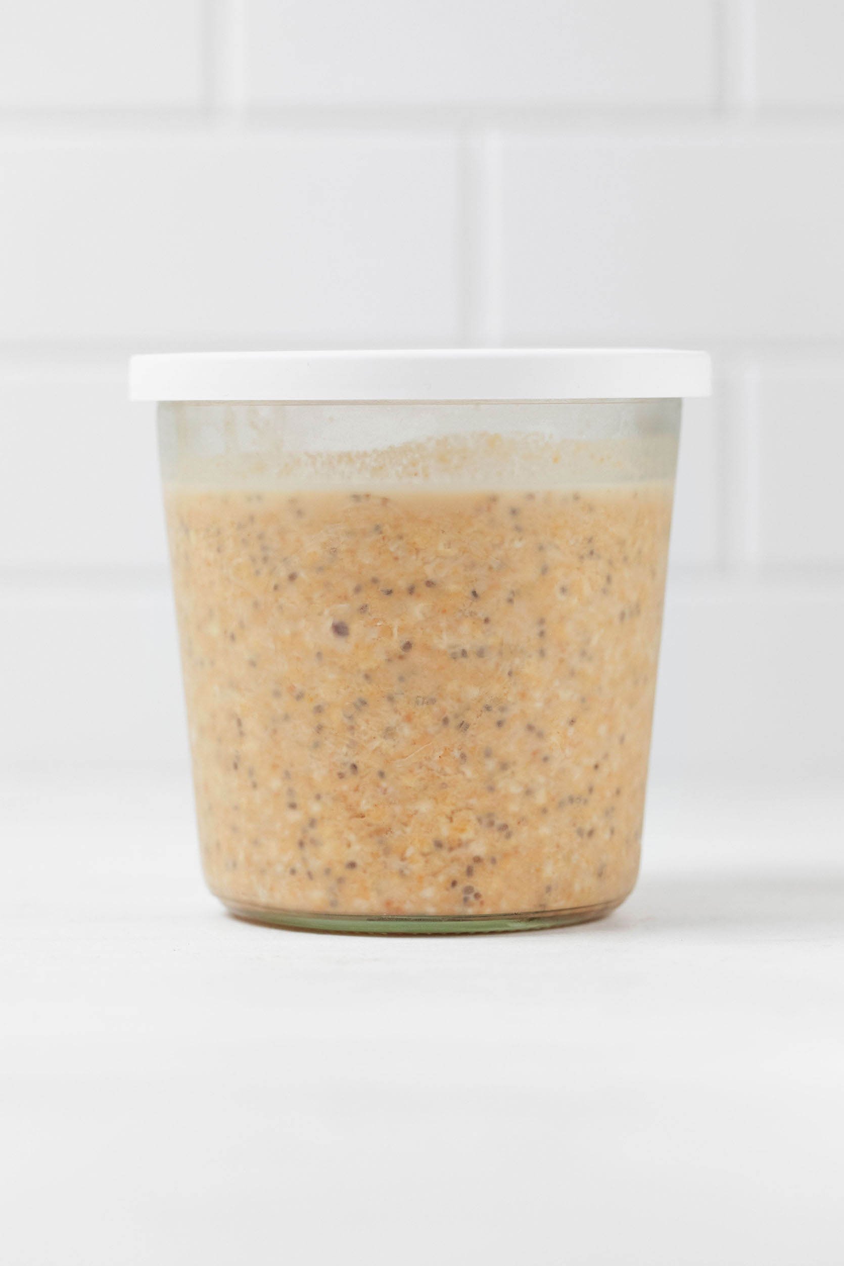A glass mason jar with a white lid is resting on a white surface. It holds a mixture of vegan pumpkin overnight oats with chia seeds.