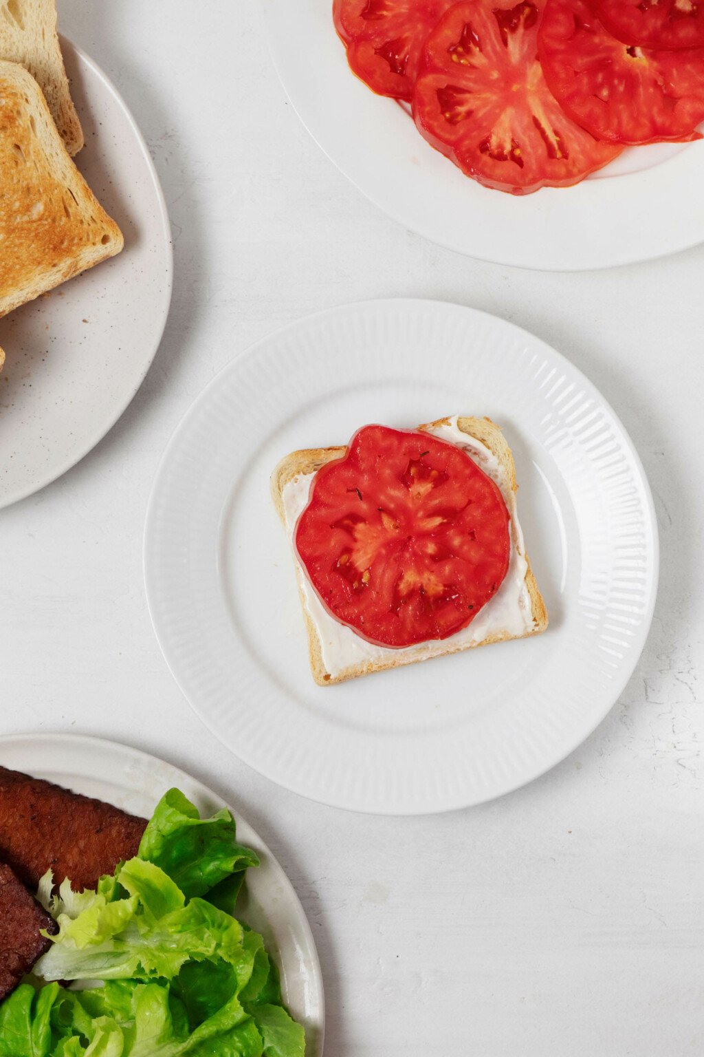 A slice of toast has been topped with a single, round slice of tomato and mayonnaise. It's served on a small white plate.