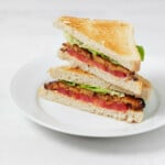 Two halves of a vegan BLT sandwich are stacked on top of each other. The rest on a round, white plate.