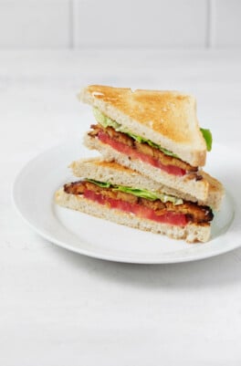 Two halves of a vegan BLT sandwich are stacked on top of each other. The rest on a round, white plate.