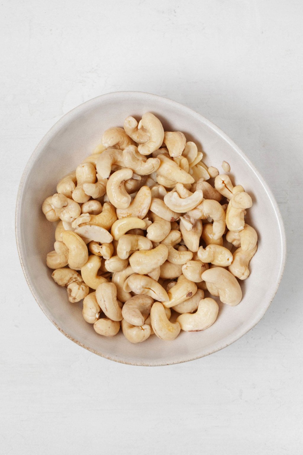 A bowl of raw cashews is resting on a white surface.
