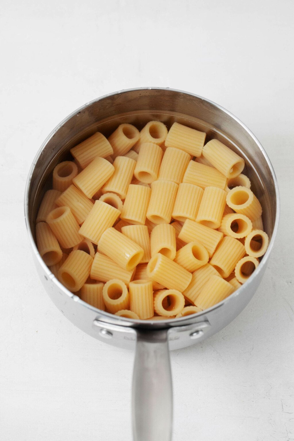 A stainless steel sauce pot is filled with cooked rigatoni. It rests on a white surface.