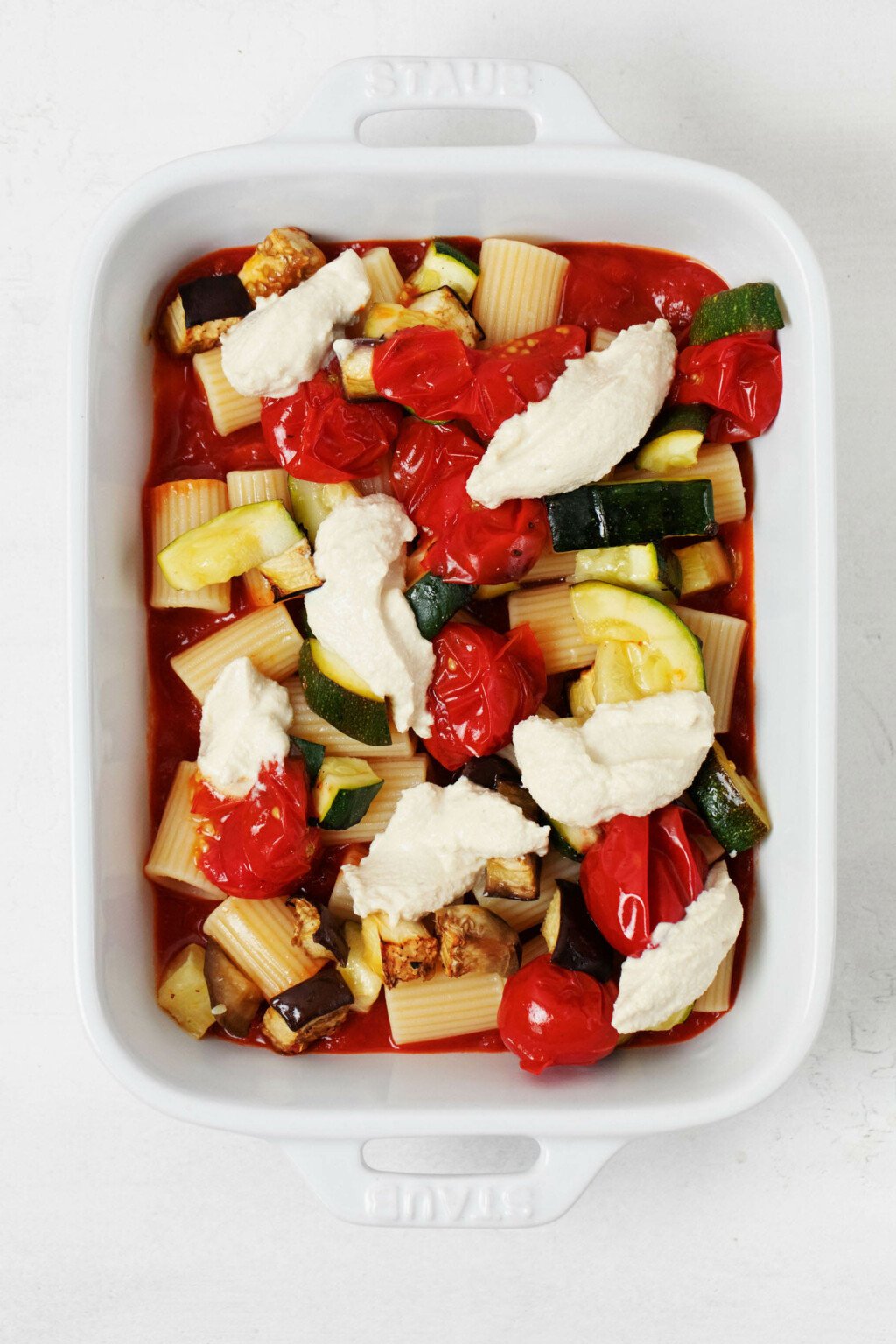 A small, white, rectangular baker has been filled with layers of roasted vegetables and pasta.