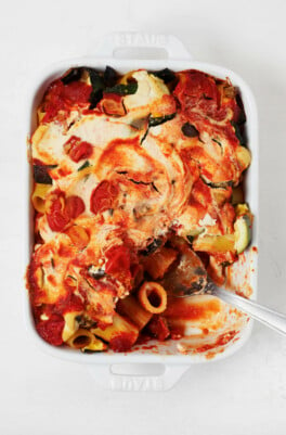 A white, rectangular baking dish holds a vegan pasta bake with ricotta and roasted summer vegetables.