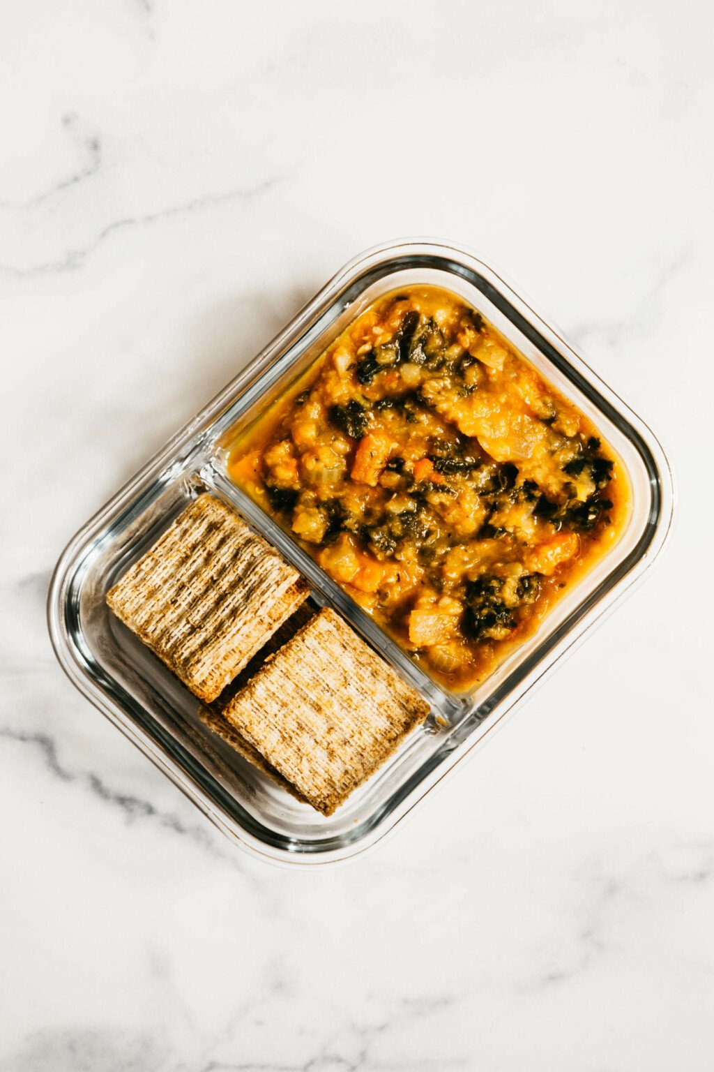 A divided glasslock container is filled with vegetable soup and whole grain crackers.