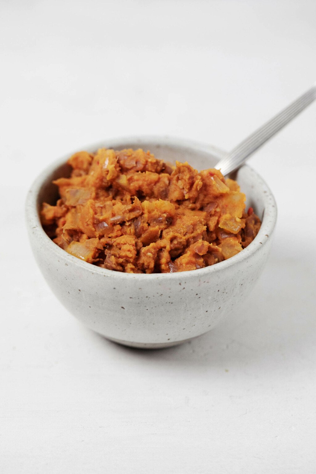 A small, round white bowl holds homemade vegan refried beans. A spoon is being used to serve them.