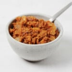 A small, round white bowl holds homemade vegan refried beans. A spoon is being used to serve them.