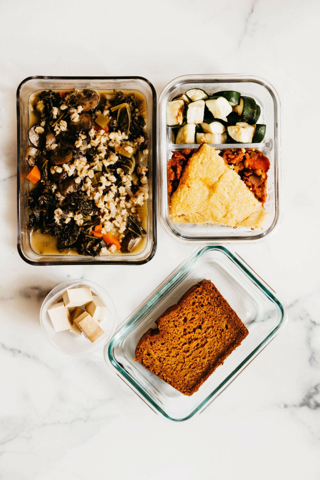 Various storage containers are resting on a white surface. They're filled with recipes that are part of a vegan meal prep.