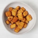 An asymmetrical, ceramic bowl holds brown tempeh "Nuggets" It is located on a white surface.