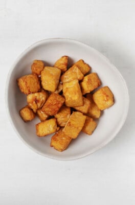 An asymmetrical, ceramic bowl holds browned tempeh "nuggets." It rests on a white surface.