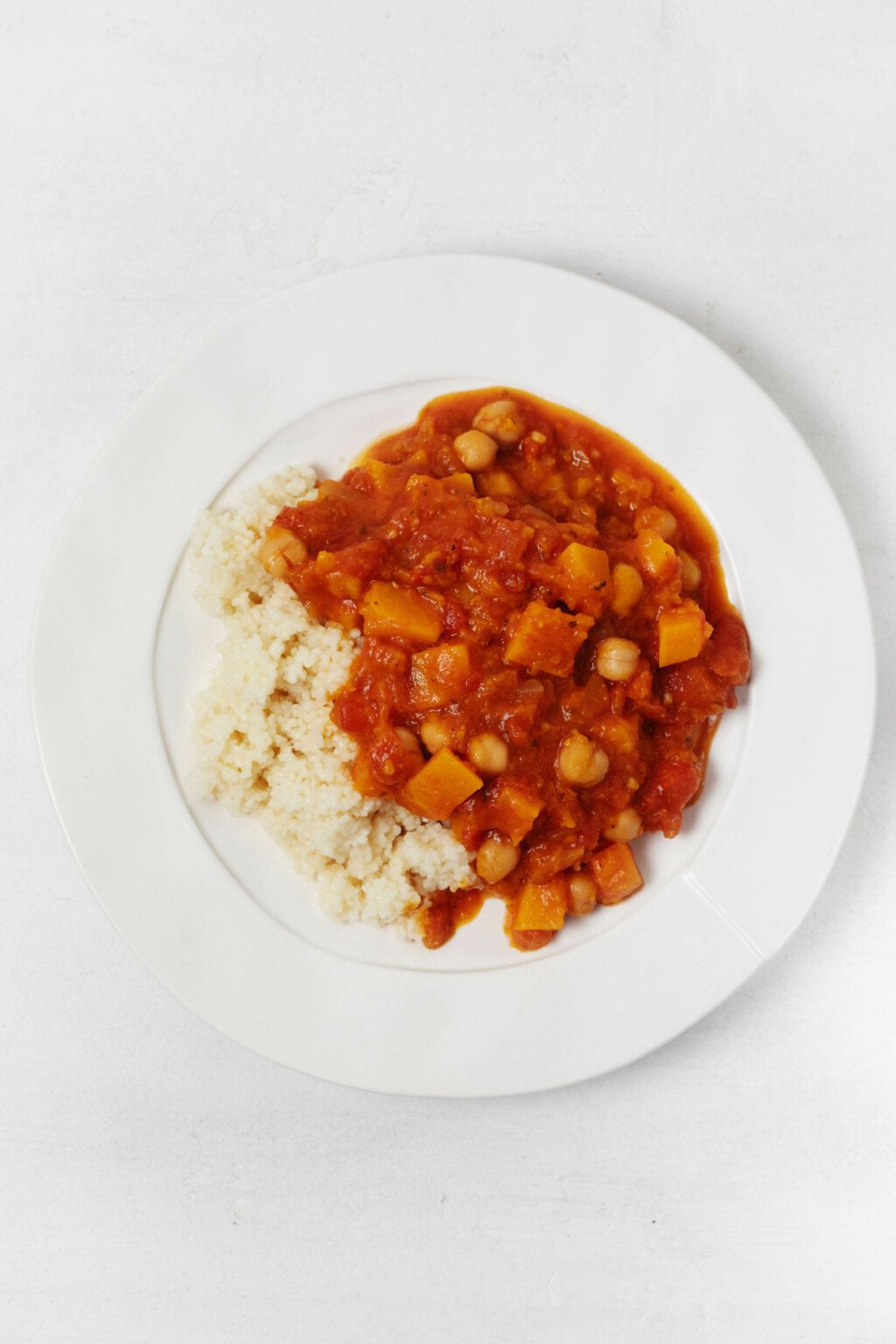 A large, rimmed white plate is covered in couscous and a Moroccan-inspired, butternut chickpea stew.