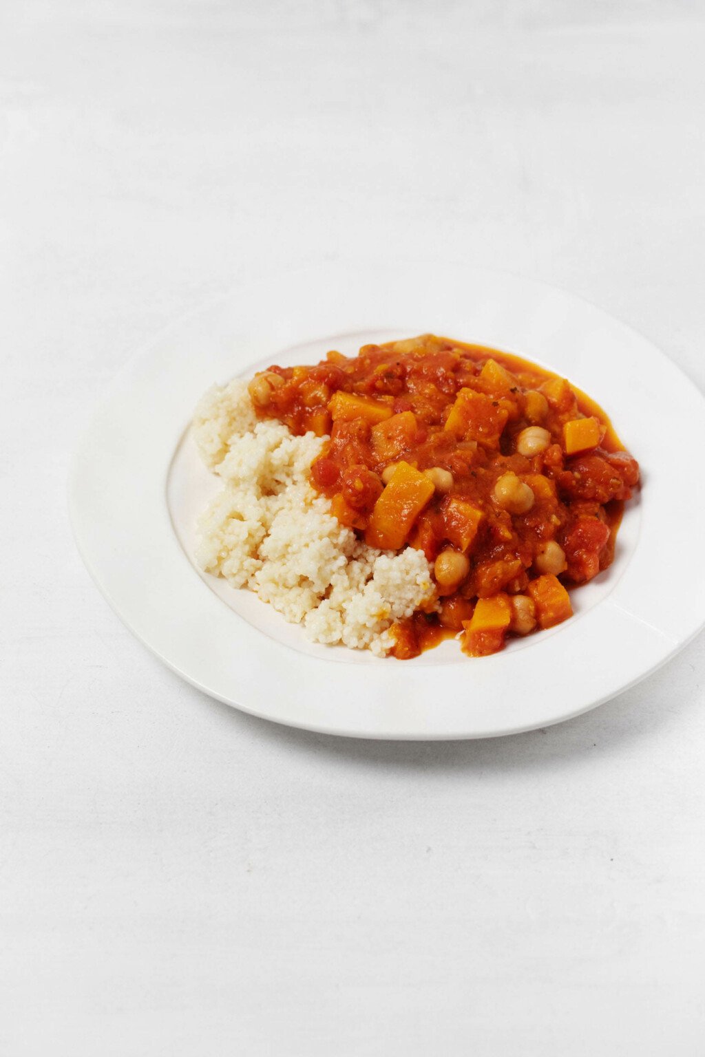 A round, rimmed white plate rests on a white surface. On the plate is couscous and a Moroccan-inspired, butternut chickpea stew.
