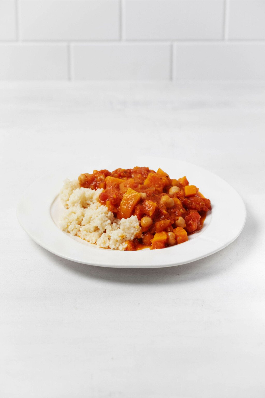 A rimmed, round white plate rests on a white surface. It's covered with a mixture of tomatoes, squash, and chickpeas.