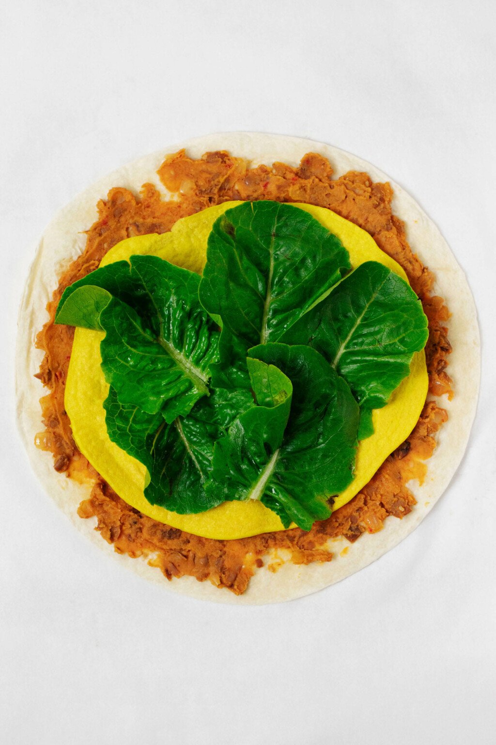 A round, flour tortilla has been layered with refried beans, a vegan egg mixture, and romaine lettuce leaves.