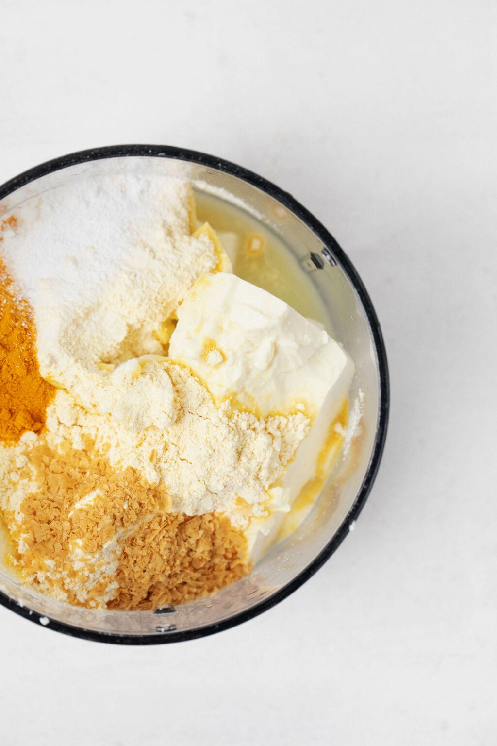 The clear bowl of a food processor holds the components for a homemade, liquid vegan egg, including tofu and flour and spices.