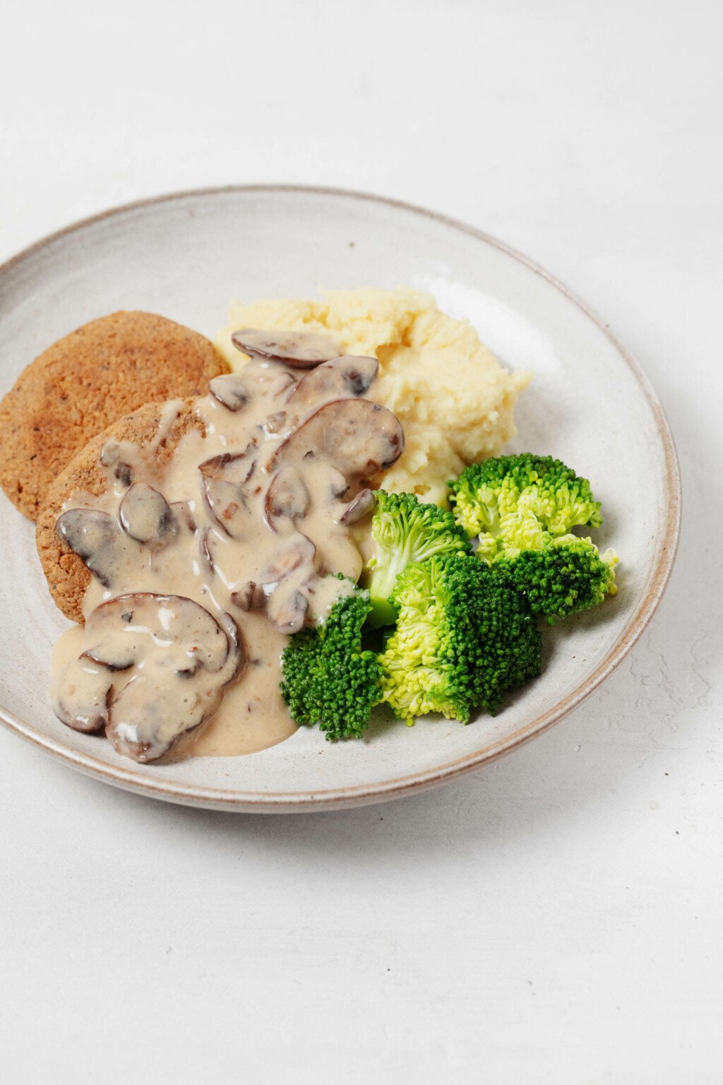 A tempeh-based version of vegan "salisbury steak" has been plated in a round, white dish, with broccoli, mushroom gravy, and mashed potatoes.