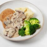 A tempeh-based version of vegan "salisbury steak" has been plated in a round, white dish, with broccoli, mushroom gravy, and mashed potatoes.