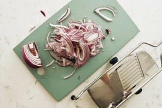 Thinly sliced red onions are laid out on a cutting board with a silver mandolin slicer nearby.