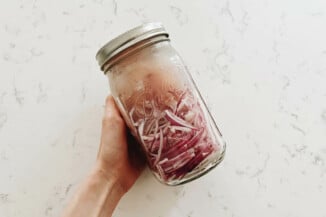 A large, ball mason jar contains pickled vegetables. Someone is holding it in her hand against a white surface.