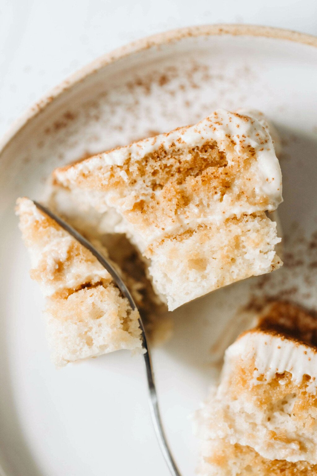A fluffy and creamy slice of vegan tiramisu has been dusted with cocoa. It's being cut with a fork.