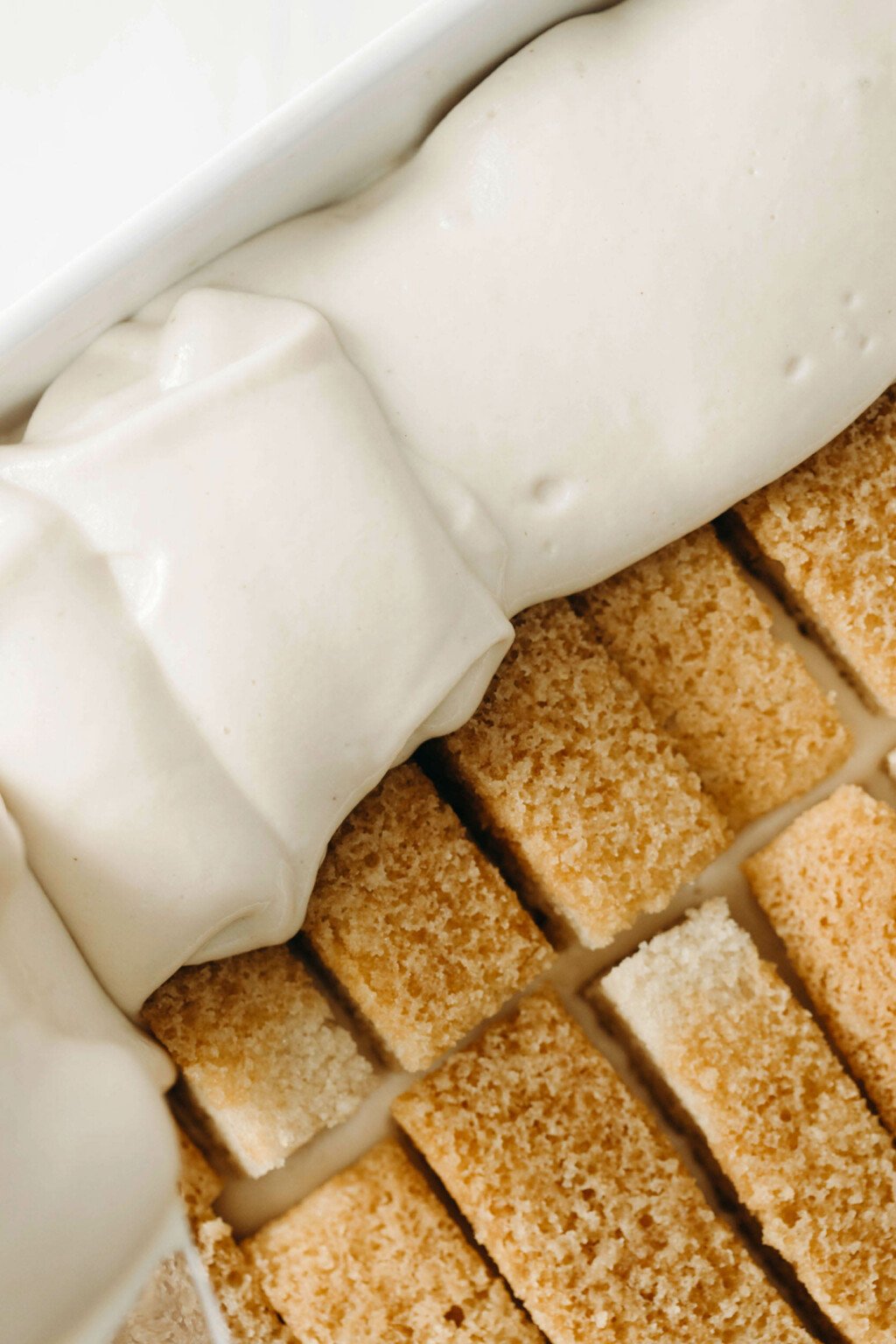 Rectangular slices of vanilla cake are being topped with a layer of a rich, creamy white mixture.
