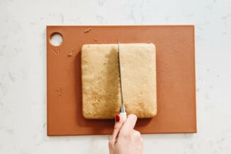 A square vanilla cake is being cut in half on a cutting board.