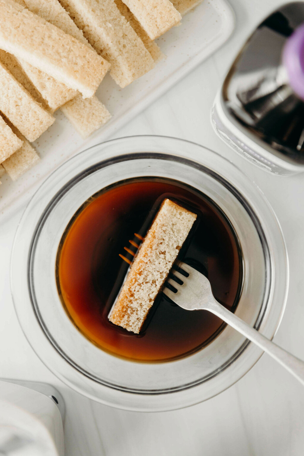 A rectangular slice of cake is being dipped into a mixture of coffee and liqueur. A fork suspends the cake in the liquid.