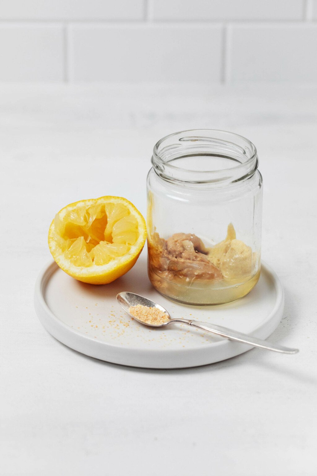 A jar with salad dressing ingredients, half a lemon and a spoonful of mustard resting on a round white plate.