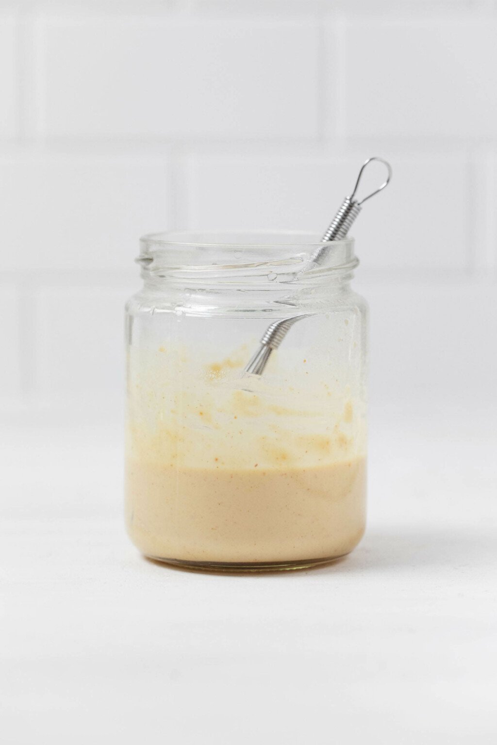 A jar of lemon tahini dressing and a small whisk are resting on a white surface.