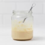 A jar of lemon tahini dressing and a small whisk are resting on a white surface.