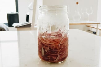 A mason jar is resting on a white countertop with a shiny finish. It holds freshly pickled red onions.