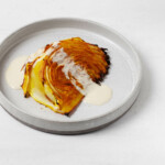 A wedge of spice-roasted cabbage steak is resting on a small, round plate, drizzled with some white creamy sauce.