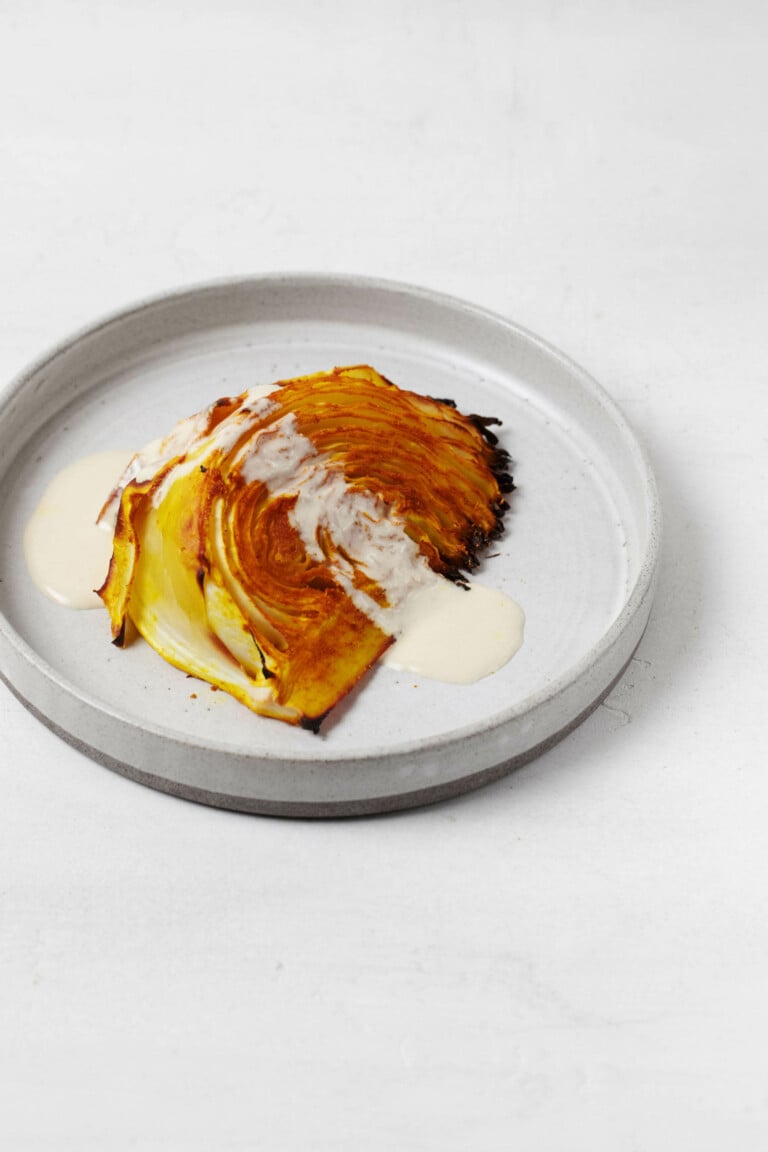 A wedge of spice-roasted cabbage steak is resting on a small, round plate, drizzled with some white creamy sauce.