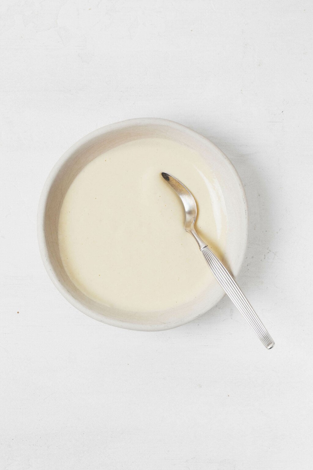 Ένα μικρό, λευκό μπολ με ντρέσινγκ ταχίνι λεμόνι Ντιζόν στηρίζεται σε μια λευκή επιφάνεια.