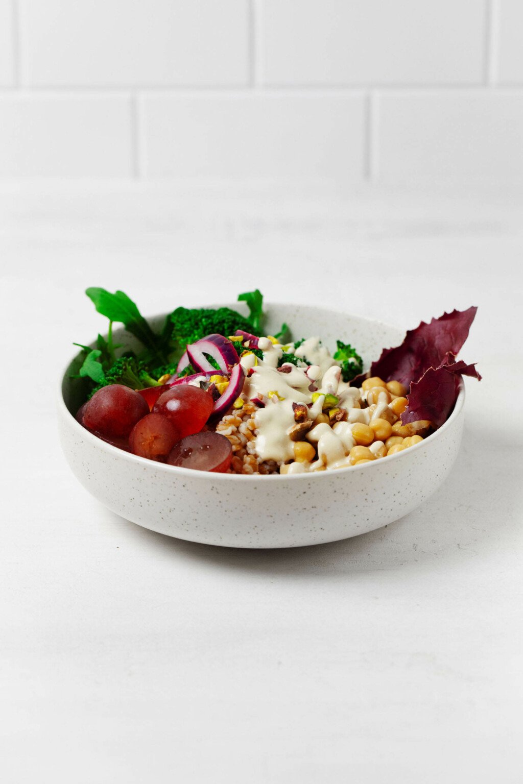 An angular image of a white bowl, filled with farro and other plant-based ingredients, resting on a white background.