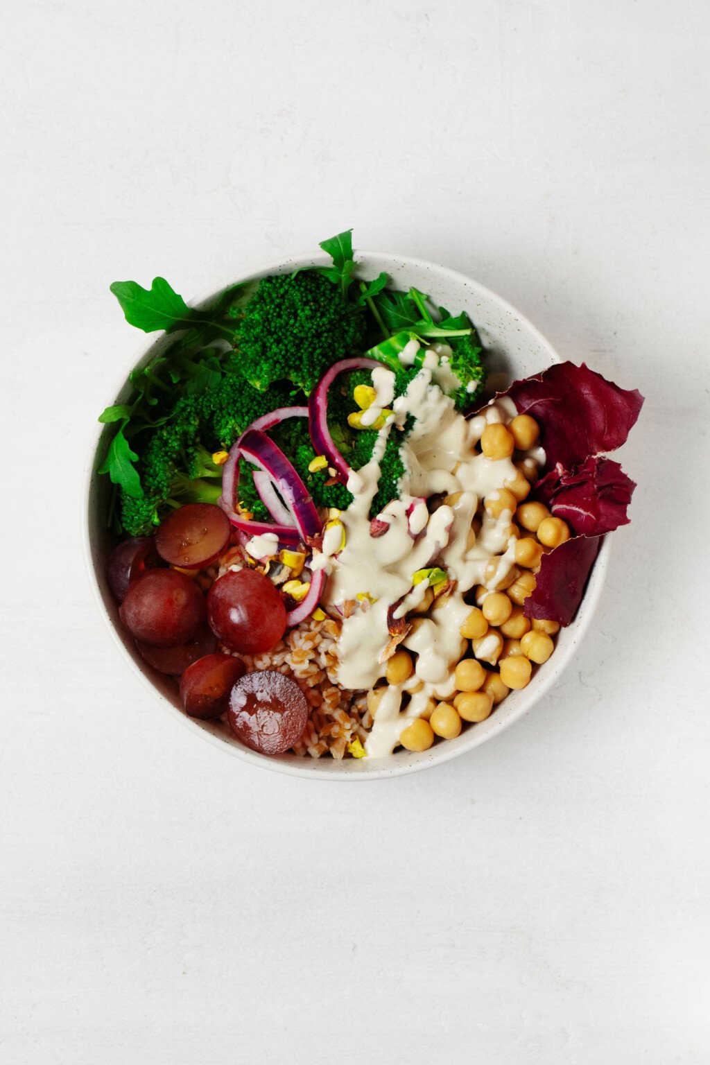 An overhead image of a white, round ceramic dish that contains the ingredients for a vegan farro bowl. It's topped with tahini dressing and chopped, shelled pistachios.