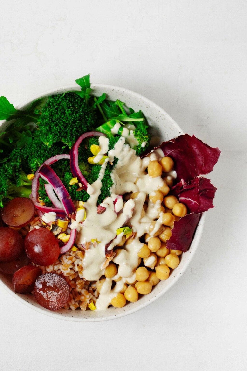 An overhead image of a white bowl that is filled with farro, chickpeas, pickled onions, and a drizzle of tahini dressing.