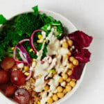 An overhead image of a white bowl that is filled with farro, chickpeas, pickled onions, and a drizzle of tahini dressing.