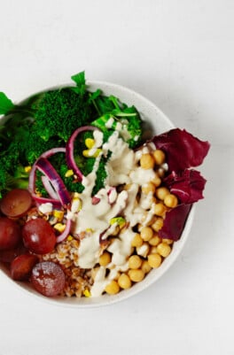 An overhead image of a white bowl that is filled with farro, chickpeas, pickled onions, and a drizzle of tahini dressing.