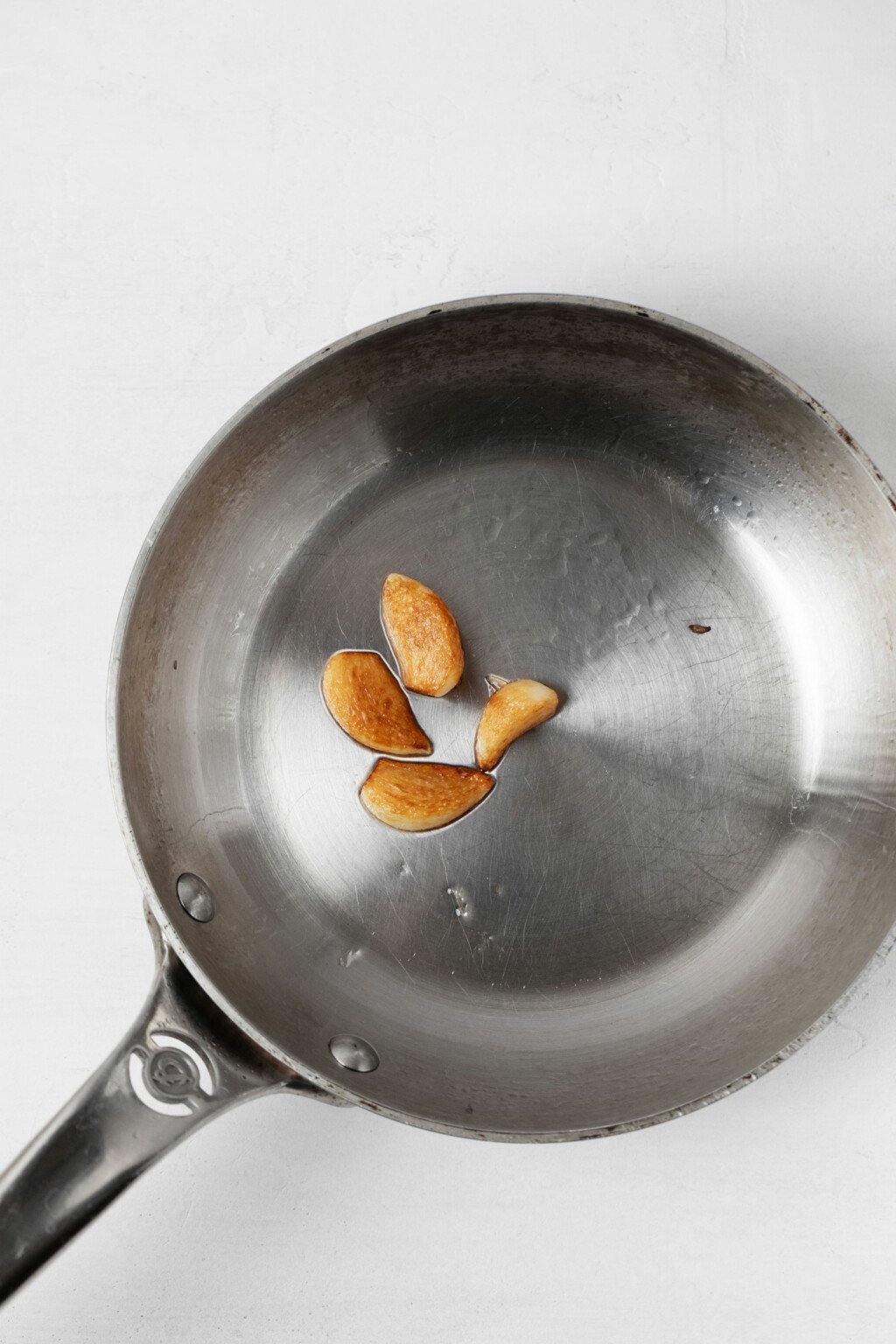 Four cloves of garlic are being browned in a small, metal frying pan.