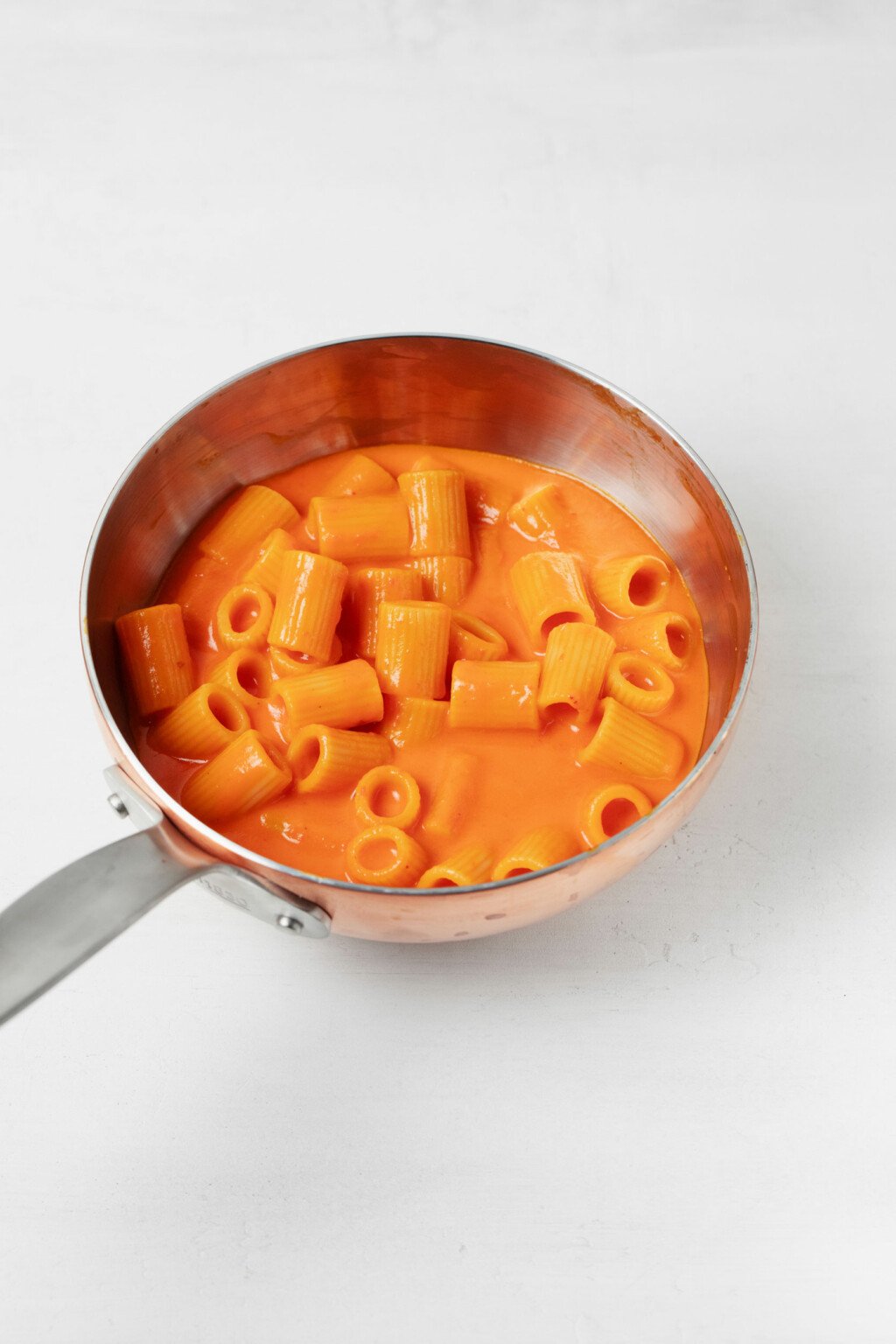 A small, stainless steel frying pan is being used to warm up a red pepper pasta with creamy sauce.