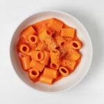 A creamy, vegan red pepper pasta is served in a round, white bowl. The bowl rests on a white surface.