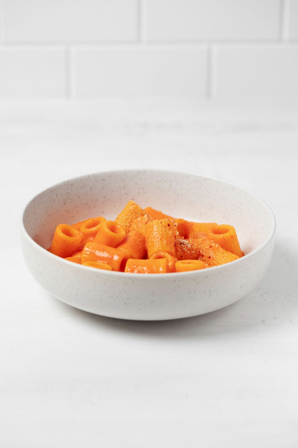 A shallow, round white bowl holds rigatoni pasta with a creamy, red-colored sauce. It rests on a white surface.