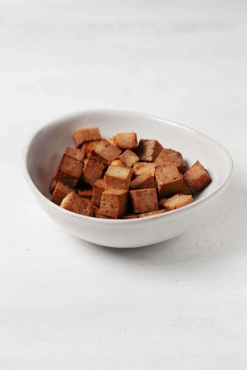 An asymmetrical white bowl holds brown-colored cubes of teriyaki tofu.