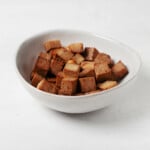 An asymmetrical white bowl holds brown-colored cubes of teriyaki tofu.