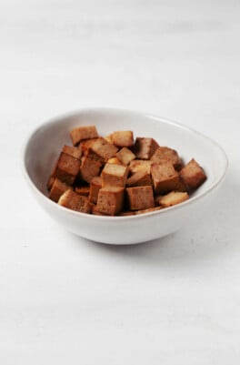 An asymmetrical white bowl holds brown-colored cubes of teriyaki tofu.